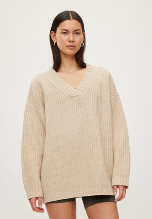 Femme portant un pull beige ample en maille à col en V et un short foncé, debout devant un fond clair uni.