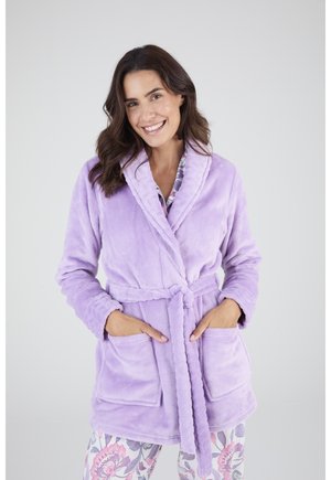 Femme souriante portant un peignoir lavande doux noué à la taille, les mains dans les poches avant et un pantalon de pyjama fleuri.