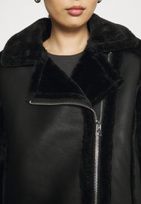 Kvinna i svart läderjacka med fluffig krage och silverdragkedja, läpparna synliga, ljus hudton, neutral bakgrund.