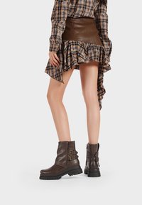 Stivali alla caviglia in pelle marrone con finitura testurizzata, suole spesse e dettagli a doppia fibbia. Abbinati a una camicia a quadri e una gonna con volant.