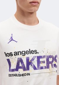 Κοντινό πλάνο ενός ατόμου που φοράει λευκό πουκάμισο με μωβ και μαύρο κείμενο "Los Angeles Lakers" και μωβ λογότυπο Jumpman στο πάνω μέρος του στήθους.