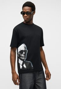 Νεαρός άνδρας που φοράει μαύρα γυαλιά ηλίου, μαύρο t-shirt με εκτυπωμένο ασπρόμαυρο πορτρέτο και σκούρα τζιν, μπροστά σε απλό φόντο.