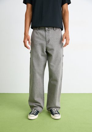 Graue Cargo-Hosen mit lockerer Passform, seitlichen Taschen, geradem Beinschnitt und vorderem Knopfverschluss. Getragen mit schwarzen Sneakers.