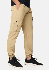 Pantalones cargo beige con puños elásticos, dos bolsillos laterales con botón y un corte relajado, combinados con zapatillas deportivas negras.