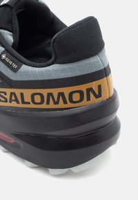 Čierne a šedé športové tenisky s textúrovaným textilným zvrškom, GORE-TEX brandingom, robustnou čiernou podrážkou a výrazným oranžovým logom.