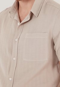 Camicia beige a maniche corte in tessuto testurizzato, con tasca cucita, chiusura a bottoni e dettagli del colletto piccoli.