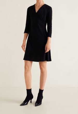 Kobieta ubrana w czarną sukienkę do kolan z rękawami do łokci i dekoltem w serek, do tego czarne botki na obcasie wykonane w stylu skarpetkowym.