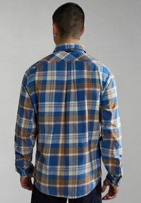 Hombre vistiendo una camisa de cuadros azul, blanca y marrón de manga larga, visto desde la parte posterior contra un fondo gris liso.