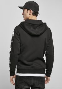 Sort hoodie med hætte, prydet med hvide grafiske designs på ærmerne. Modellen bærer en sort kasket og mørke jeans, hvilket giver et afslappet look.
