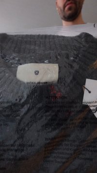 Γκρι πουλόβερ συσκευασμένο σε πλαστική σακούλα με μια λευκή ετικέτα που φέρει την επωνυμία «Jack & Jones» και το μέγεθος «XL». Διακρίνονται χαρακτηριστικές λεπτομέρειες υφής και υλικού.