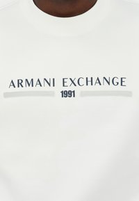 Chemise blanche avec le texte « ARMANI EXCHANGE » en lettres majuscules noires et « 1991 » centré en dessous entre deux lignes grises horizontales.