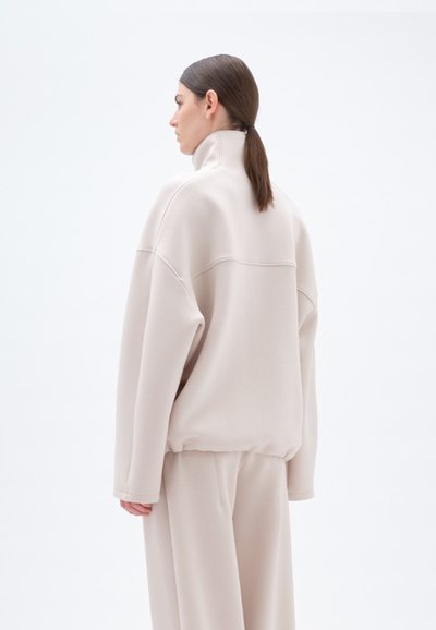 Lys beige sweatshirt med høy krage, avslappet passform, droppede skuldre og ribbestrikket hem, parret med matchende bunnplagg, strukturert stoff.