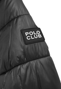 Svart vadderad jacka med en slät, glänsande yta och en upphöjd svart lapp med orden "POLO CLUB" i vitt.