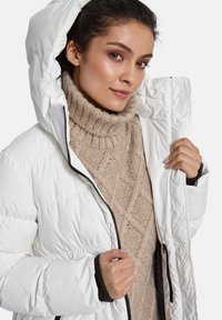 Vrouw in een beige kabelgebreide coltrui onder een witte gewatteerde jas met capuchon, die de jas met één hand openhoudt.