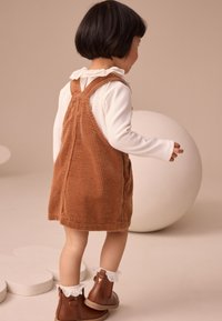 Vestito di velluto a coste marrone con bretelle, indossato sopra una camicia bianca a maniche lunghe, abbinato a stivaletti marroni con dettagli in pizzo.