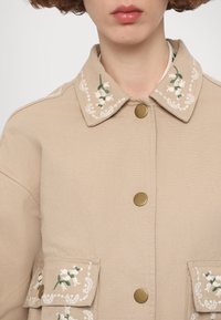 Beige canvas jacket with embroidered floral design on collar and pocket flaps, featuring gold-tone buttons and a textured finish.