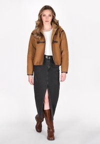 Bruine shearling-voering jas met bruine accenten, witte top, zwarte spijkerrok met een voorknip en bruine kniehoge laarzen.