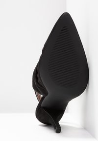 Scarpa nera in suede con tacco alto, punta affusolata e suola in gomma testurizzata, inclinata su uno sfondo bianco per una chiara visibilità.