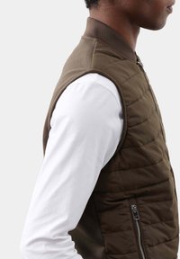 Gepolsterte ärmellose Jacke in Dunkelbraun mit einem gerippten Kragen, gesteppter Textur und einer mit Reißverschluss versehenen Seitentasche. Trägt man über einem weißen Langarmshirt.