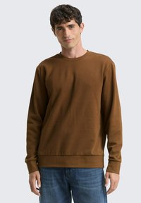 Bruine lange mouw sweatshirt met ronde halslijn, geribde manchetten en zoom, gedragen met blauwe spijkerbroek. Soepele textuur, casual design.