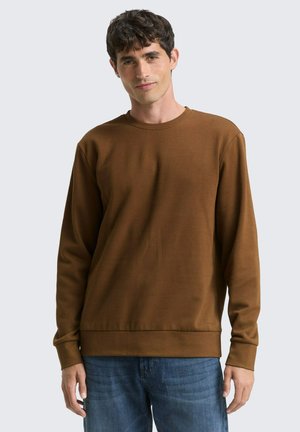 Bruine lange mouw sweatshirt met ronde halslijn, geribde manchetten en zoom, gedragen met blauwe spijkerbroek. Soepele textuur, casual design.