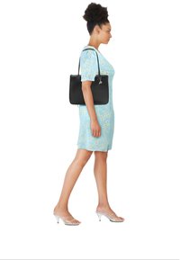 Lichtblauwe jurk met geel bloemenpatroon, zwarte draagtas, doorzichtige sandalen met hak, drager loopt in profiel.