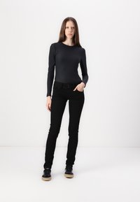 Eine Frau steht vor einem weißen Hintergrund und trägt ein enges schwarzes Langarmoberteil, schwarze Skinny-Jeans und schwarze Sneakers mit beigen Sohlen.