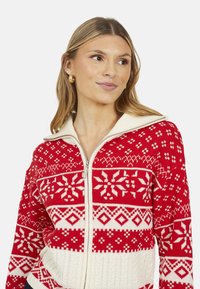 Maglione rosso e crema con zip e colletto alto. Presenta motivi a fiocco di neve e geometrici, con la parte inferiore a coste testurizzate.
