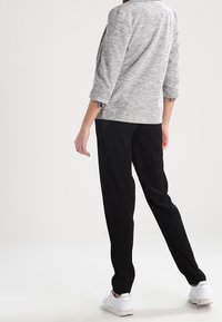 Femme portant une veste grise texturée, un pantalon noir et des baskets blanches Reebok, debout devant un fond blanc.