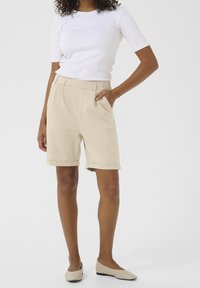 Shorts beige sur mesure avec ourlets retournés, poches latérales et texture de tissu lisse, associés à un haut blanc ajusté à manches courtes.