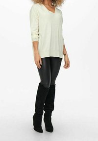 Femme portant un pull en V crème, des leggings en cuir noir et des bottes noires en suède montant jusqu'au genou, se tenant contre un fond blanc.