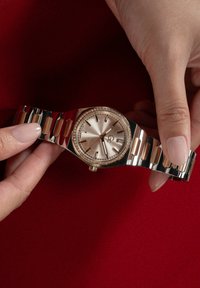 Roséguldtonad klocka med en texturerad urtavla, diamantaccentuerad bezel och länkad metallarmband. Hållen av en hand mot en röd bakgrund.