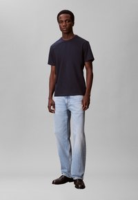 Marineblå bomuld T-shirt kombineret med lyseblå jeans med lige ben. Modellen står i sorte lædersko mod en neutral baggrund.