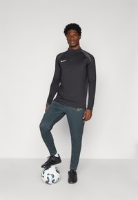 Czarna sportowa bluzka z długim rękawem z szarymi akcentami i logo Nike, w połączeniu z ciemnozielonymi spodniami sportowymi z logo Nike, stojąca na piłce nożnej.