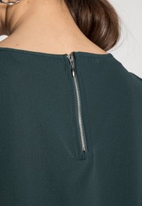 Gros plan sur l'encolure arrière d'un vêtement texturé vert foncé avec une fermeture éclair argentée partiellement zippée, porté par une personne aux cheveux bruns.