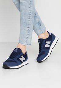 Marinblå sportskor med nät- och mockamaterial, med en vit mellansula och silverdetaljer på snörningen och logotypen.