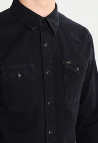 Tmavě modrá denimová košile na knoflíky s klasickým límcem, dvěma náprsními kapsami se západkami a decentním logem na vrchní kapse.