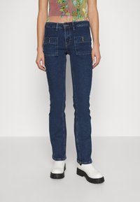En person som bär mörkblå straight-leg jeans med framknäppta fickor och vita klumpiga boots vid vristen, mot en neutral bakgrund.