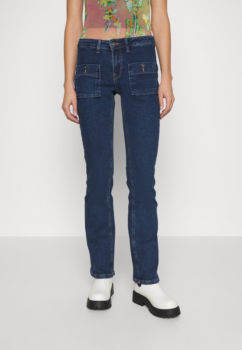 En person som bär mörkblå straight-leg jeans med framknäppta fickor och vita klumpiga boots vid vristen, mot en neutral bakgrund.