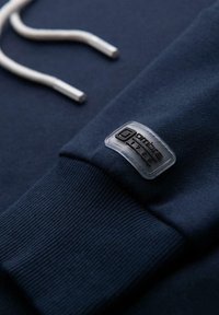 Kołnierzyk granatowego swetra z prążkowanym materiałem, białe sznurki do ściągania oraz przezroczysta łatka z napisem "ombre 1985" na rękawie.