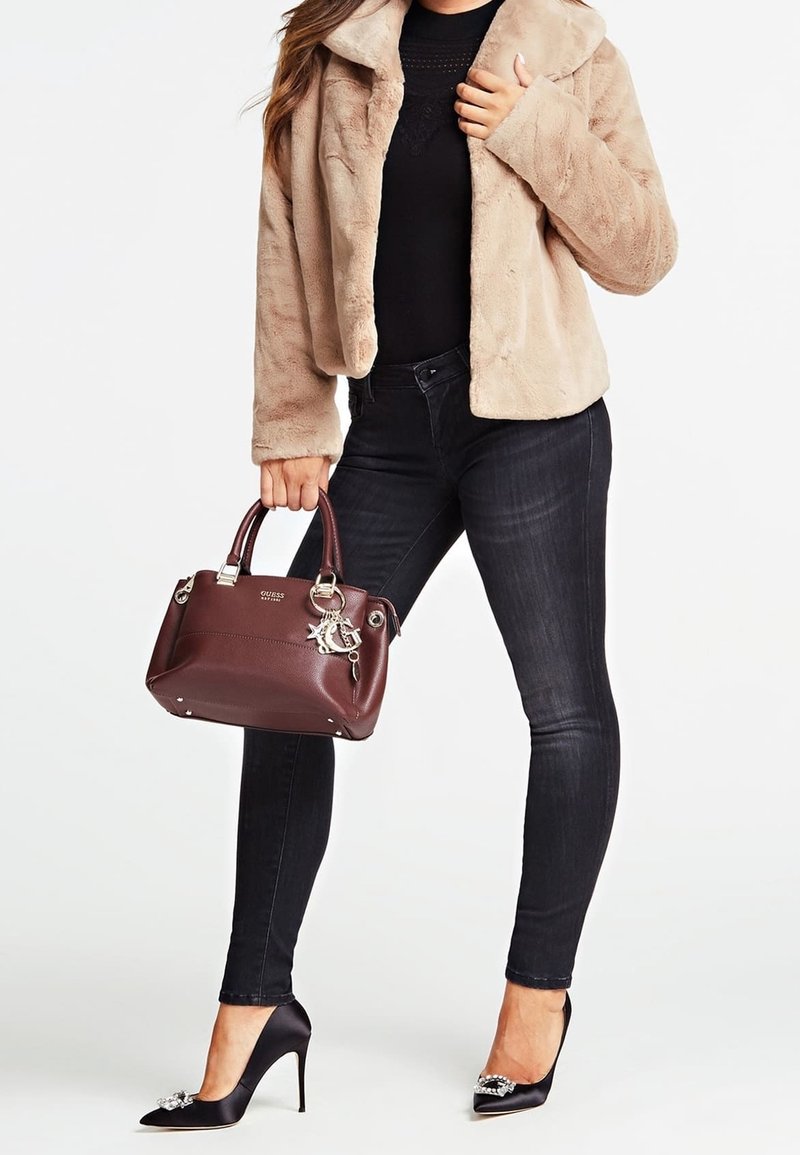 Femme portant une veste en fausse fourrure beige, un top noir, un jean skinny foncé, des talons hauts noirs décorés, tenant un sac à main en cuir marron avec un porte-clés.