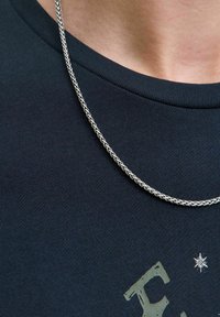 Metalen ketting met een gevlochten ontwerp, rustend op een donkerblauw katoenen t-shirt met een gestructureerde print en een klein sterdetail.