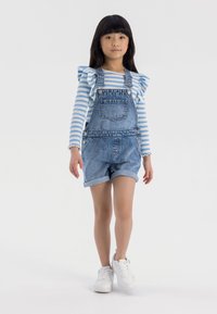 Salopette en denim avec des ourlets retroussés, portée sur une chemise à rayures bleues et blanches à manches longues avec des accents de volants. Des baskets blanches complètent le look.