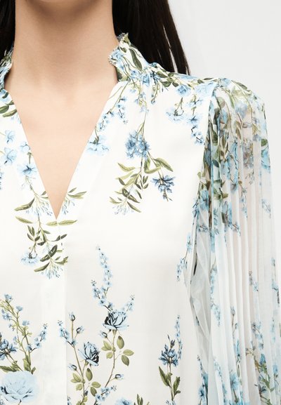 Λευκό πουκάμισο με μπλε λουλουδάτο μοτίβο, με V-neck, γιακά με βολάν και διαφανή πλισέ μακριά μανίκια με λουλουδάτο σχέδιο.