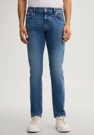 Mann trägt blaue Slim-Fit-Jeans und weiße Sneakers und steht vor einem schlichten hellen Hintergrund.