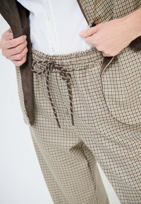 Persona che indossa pantaloni a quadri con cintura a coulisse e giacca abbinata a quadri, che sistema la giacca sopra una camicia bianca.