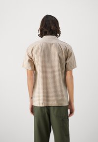 Camisa de manga corta con un patrón geométrico marrón claro, diseñada con cuello y un ajuste holgado, combinada con pantalones cargo verdes.