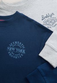 Deux sweatshirts à manches longues pliés, un bleu marine avec l'inscription "Authentic New York Brooklyn", un gris clair avec l'inscription "Bklyn New York".