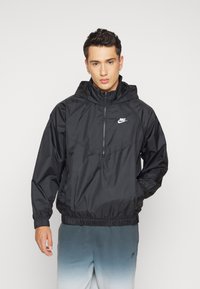 Melnā viegla Nike jaka ar kapuci, ar rāvējslēdzēju priekšā, elastīgām piedurknēm un auklu apakšmalu. Uz krūšu daļas ir neliels balts Nike logo.