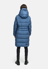 Padded blue coat with a hood, featuring stitched detailing, a cinched waist, and a logo patch, paired with black pants and boots.